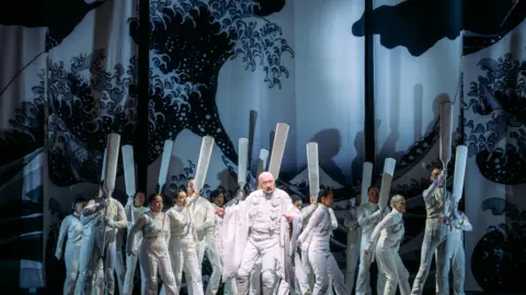 Performers on stage dressed in white with Hokusai's The Great Wave painting in the background.